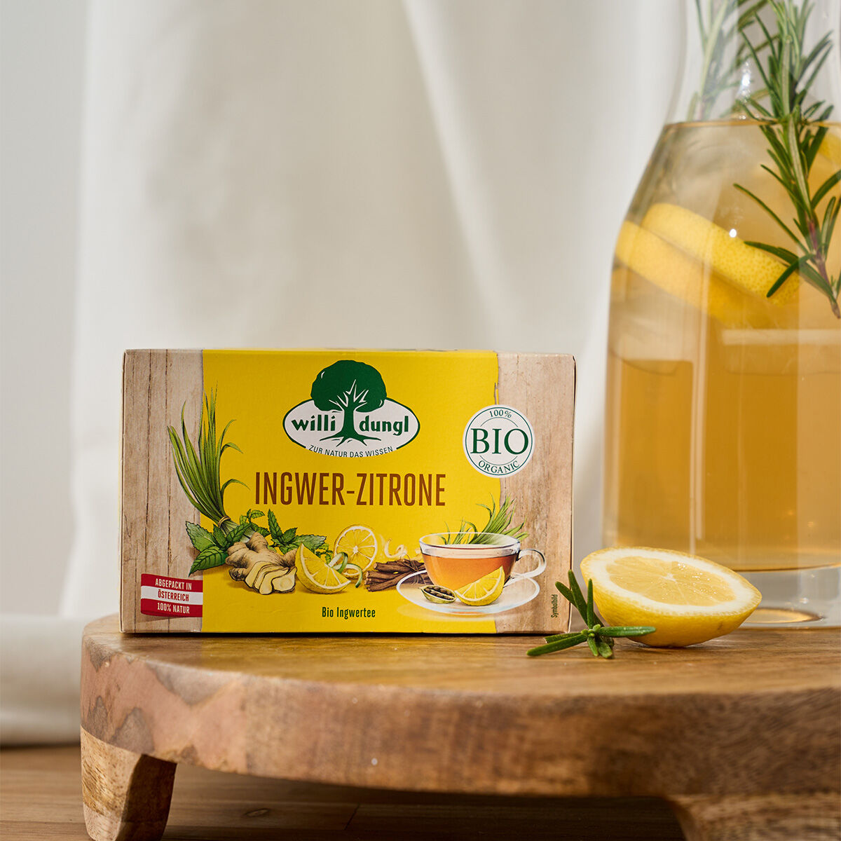 Eine Schachtel Ingwer-Zitrone BIO-Tee steht auf einem Holzbrett, im Hintergrund Zitronenscheiben, Kräuter und eine Glaskaraffe mit Zitronengetränk. Das Tageslicht sorgt für eine frische und einladende Atmosphäre.