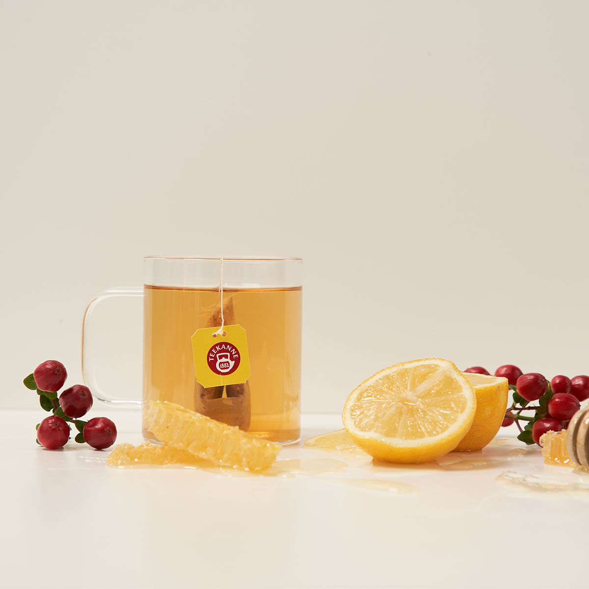 Eine klare Glastasse mit Italienischem Limone-Tee und einem Teebeutel, umgeben von Preiselbeeren, Zitronenscheiben, Honigwabe und Honig auf einer weißen Oberfläche.