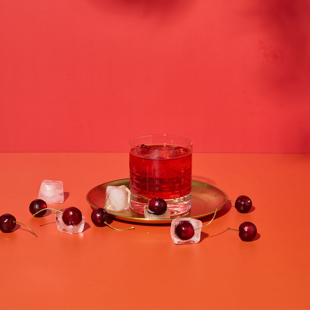 Ein Glas roter Cocktail mit Eis steht auf einem goldenen Teller, umgeben von italienischer Süßkirsche und Eiswürfeln auf einer orangefarbenen Fläche mit rotem Hintergrund – perfekt, um im Sommer La Dolce Vita zu genießen.