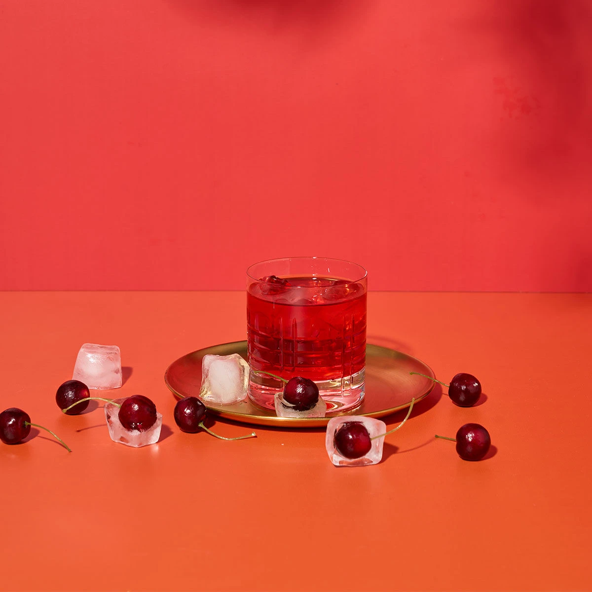 Ein Glas roter Cocktail mit Eis steht auf einem goldenen Teller, umgeben von italienischer Süßkirsche und Eiswürfeln auf einer orangefarbenen Fläche mit rotem Hintergrund – perfekt, um im Sommer La Dolce Vita zu genießen.