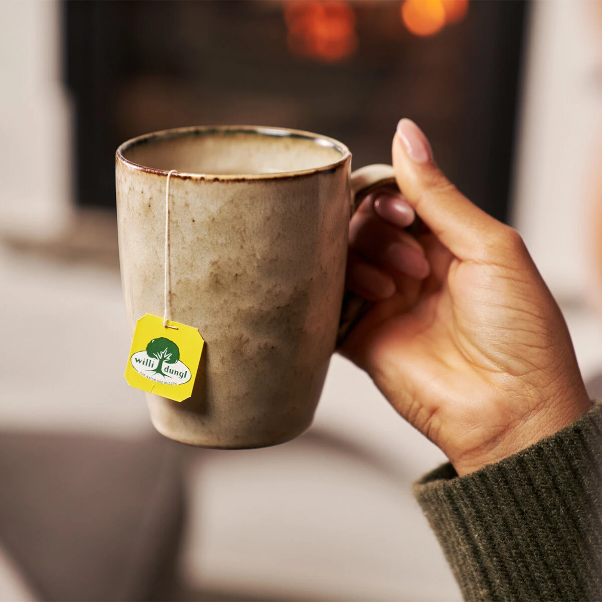 Eine Person in einem grünen Pullover hält eine Keramiktasse mit einem gelben Etikett mit einem Kamille BIO-Teebeutel und entspannt sich vor einem gemütlichen, verschwommenen Kaminfeuer im Hintergrund.