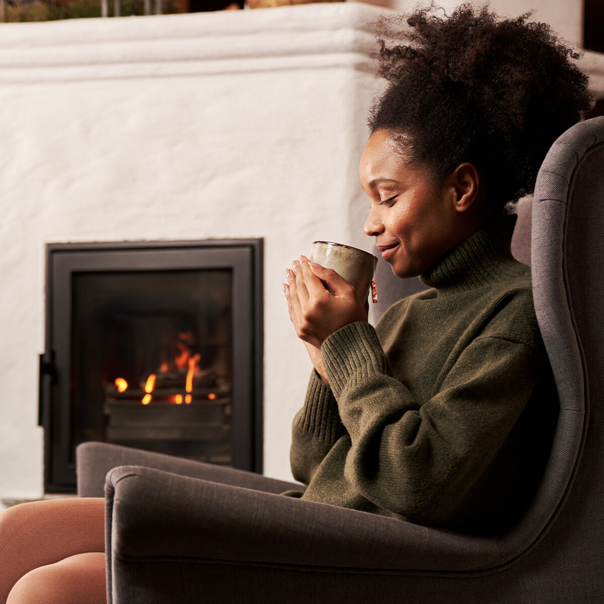 Eine Frau im grünen Pullover sitzt zufrieden in einem gemütlichen Sessel und hält ihre Wintertee-Tasse in der Hand, während im Hintergrund die Wärme von Kaminzauber aus dem Kaminfeuer glüht.