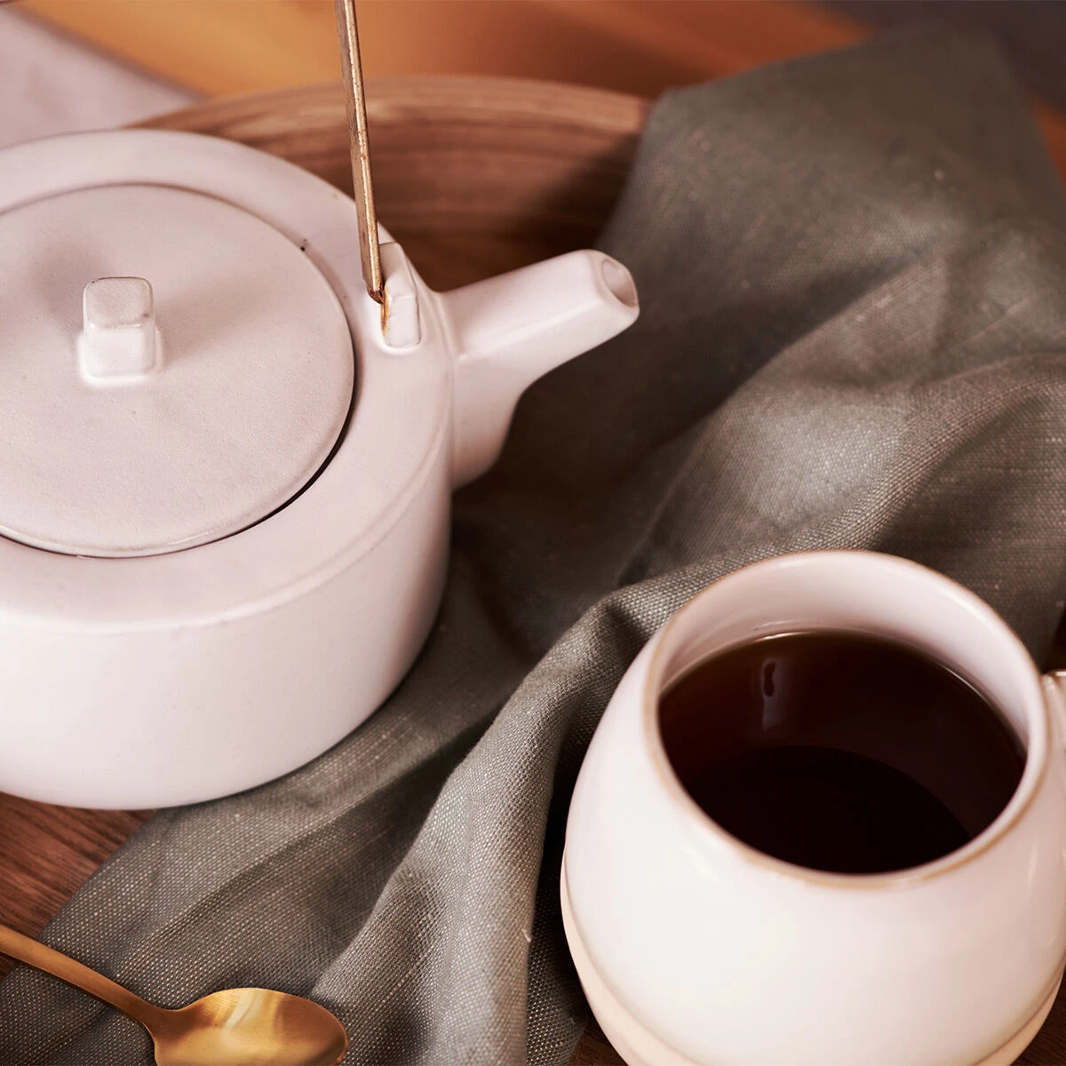 Eine weiße Teekanne und eine Tasse aus Keramik, gefüllt mit Selected. Highland Green Tea Bio Loser Tee (200 g), stehen auf einem Holztablett, begleitet von einem goldenen Löffel und einem grauen Tuch.