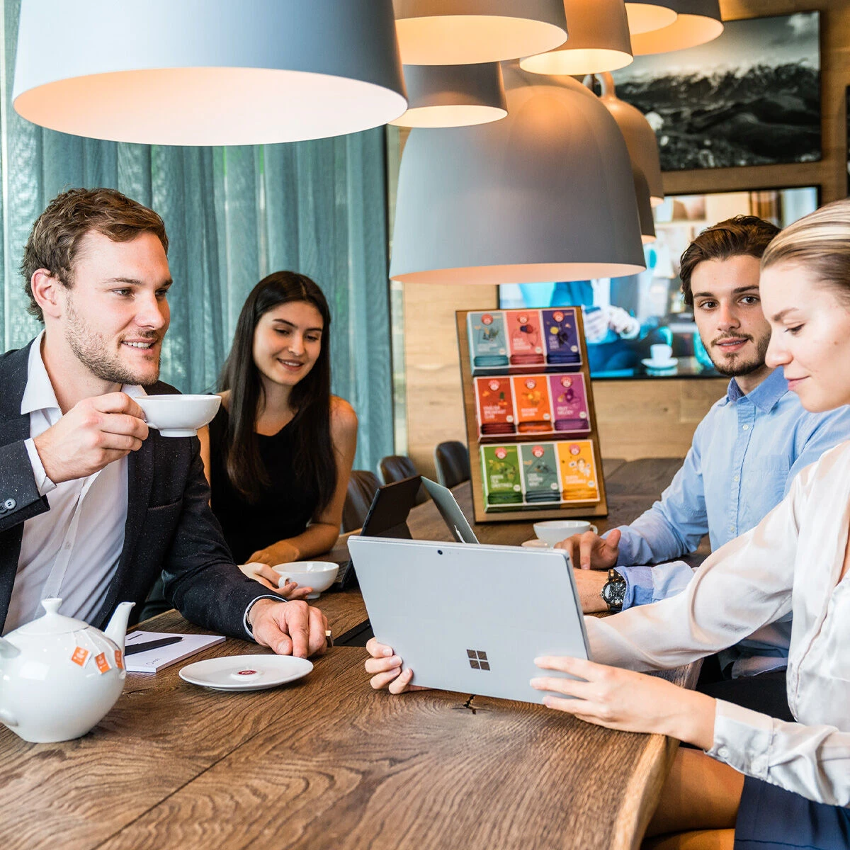 Vier junge Erwachsene treffen sich in einem modernen Café an einem Holztisch. Ein Mann genießt einen Cool Peppermint Luxury Cup BIO-Tee, während eine Frau an ihrem Laptop arbeitet. Der Tisch ist mit Teetassen, einer Teekanne und bunten Menükarten ausgesta