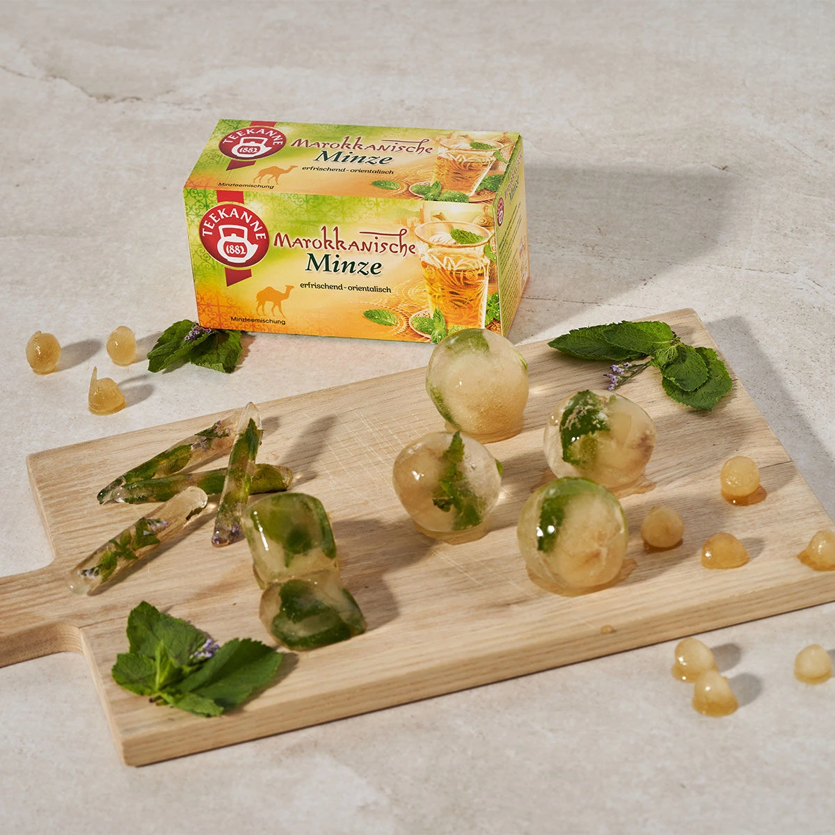 Eine Schachtel Marokkanische Minze steht hinter einem Holzbrett mit runden und dreieckigen Eiswürfeln mit Minzblättern. Loser Marokkanischer Minze-Tee und frische Minzblätter sind auf dem Brett und der Oberfläche verstreut.