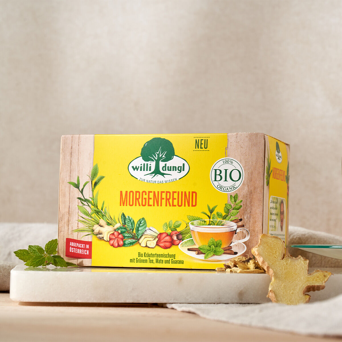 Eine Schachtel Morgenfreund Bio-Kräutertee steht auf einer Marmorplatte, daneben Minzblätter und Ingwer. Die Verpackung zeigt Kräuterbilder, eine Teetasse sowie die Etiketten „BIO“ und „NEU“.