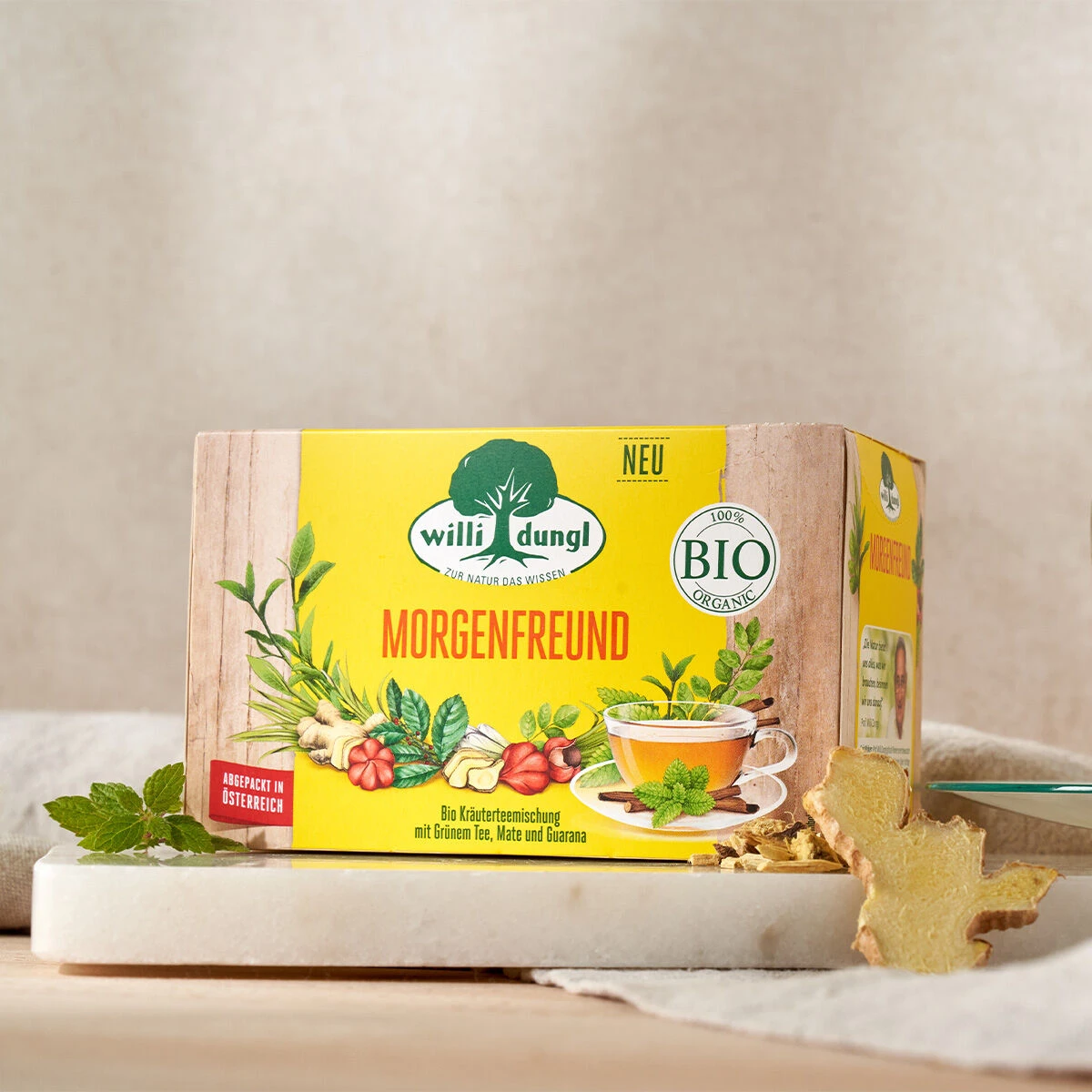 Eine Schachtel Morgenfreund Bio-Kräutertee steht auf einer Marmorplatte, daneben Minzblätter und Ingwer. Die Verpackung zeigt Kräuterbilder, eine Teetasse sowie die Etiketten „BIO“ und „NEU“.
