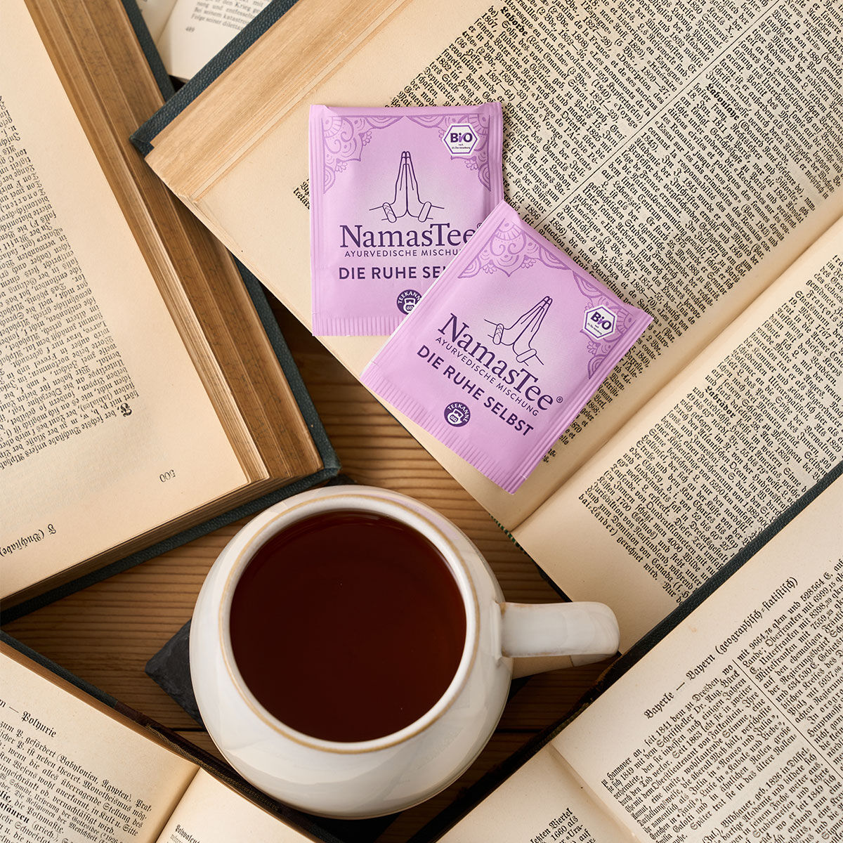 Eine Tasse Tee steht auf einem offenen Buch, umgeben von anderen. Zwei lila NamasTee BIO® Die Ruhe selbst-Sachets liegen daneben und sorgen für eine gemütliche Lese- und Teeatmosphäre.