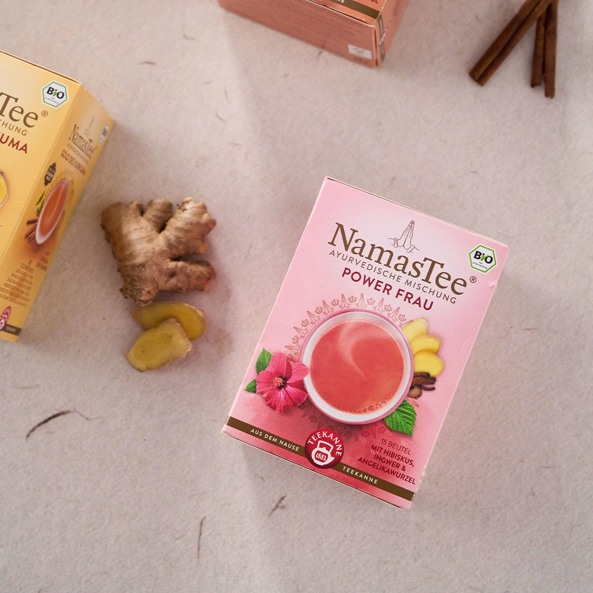 Eine rosa Schachtel mit BIO NamasTee® Power Frau Ayurveda-Tee steht auf einer hellen Oberfläche neben geschnittenem Ingwer und einer weiteren Teeschachtel. In der oberen rechten Ecke sind Zimtstangen zu sehen.
