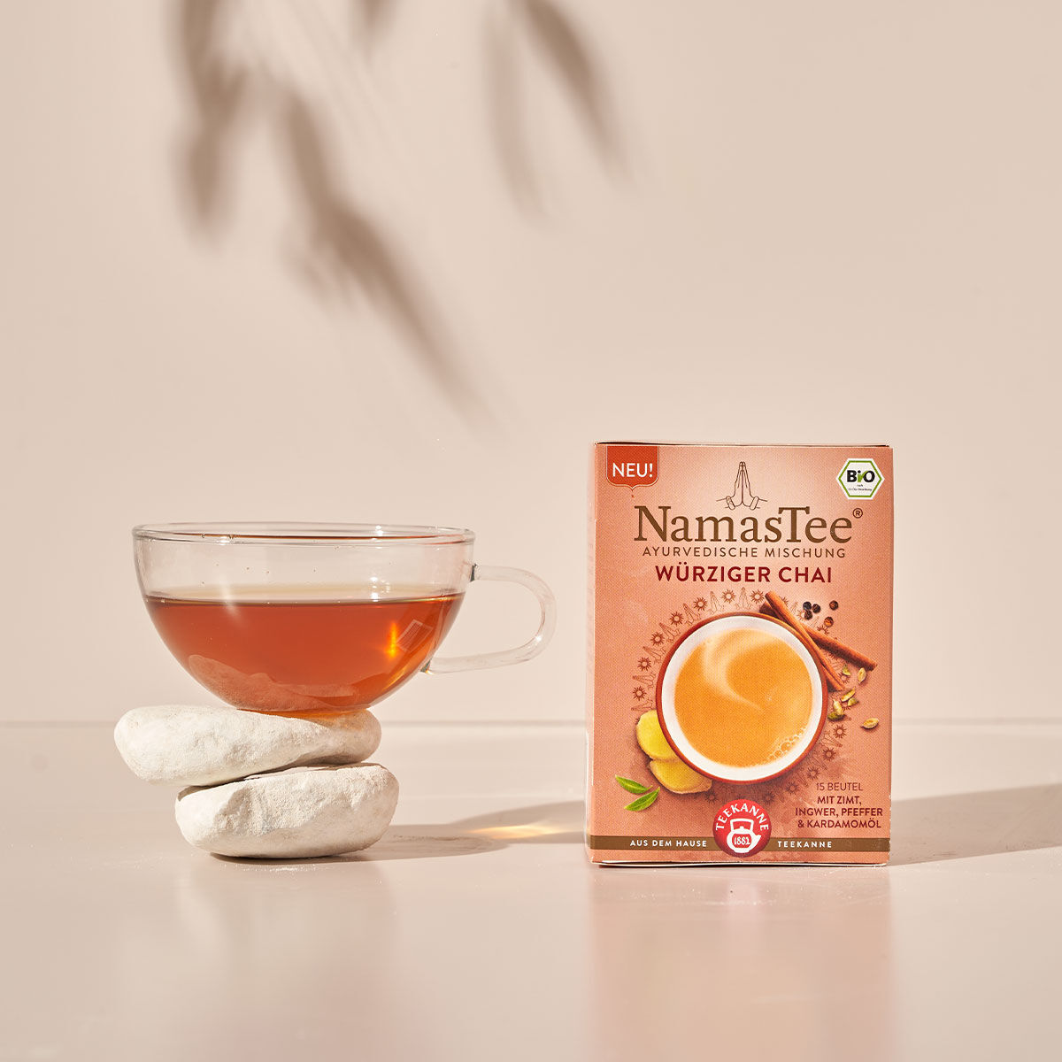 Eine Tasse Tee aus Glas steht auf gestapelten weißen Steinen neben einer Schachtel NamasTee® BIO Würziger Chai, vor einem hellbeigen Hintergrund mit weichen Schatten.