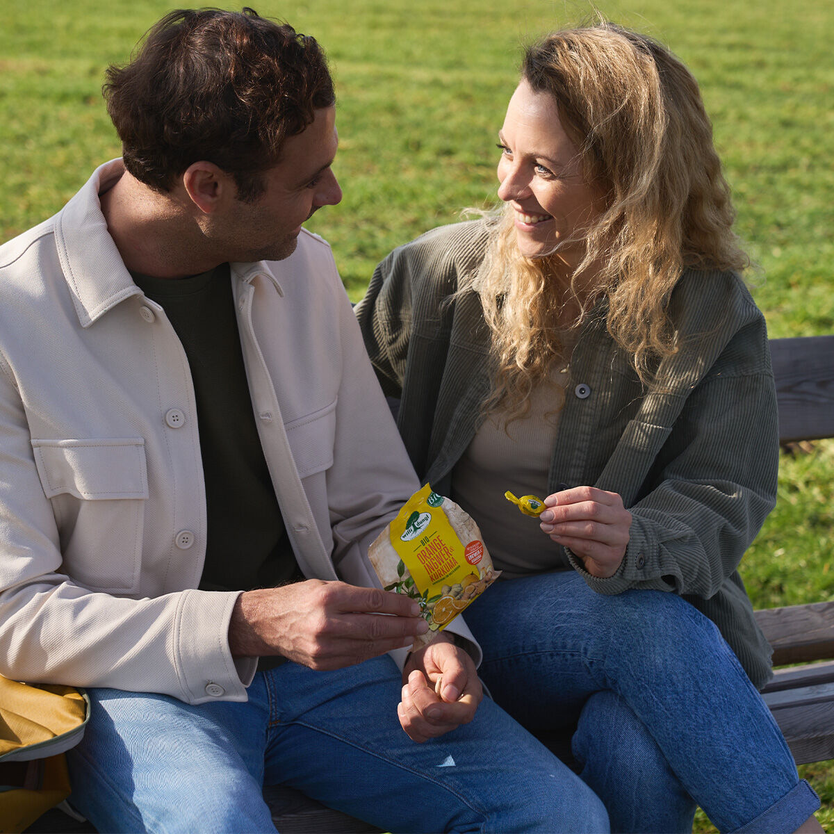 Ein Mann und eine Frau lächeln sich auf einer Parkbank an. Die Frau hält eine 65-g-Packung Orange Ingwer & Kurkuma und ein Bonbon in der Hand, während der Mann die Packung offen hält. Beide wirken entspannt und glücklich im Freien.