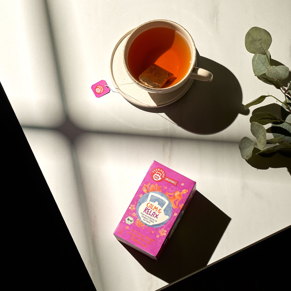 Eine Tasse Tee mit einem Teebeutel auf einer Untertasse steht auf einer weißen Fläche neben einer leuchtend rosa ORGANICS Calm & Relax BIO-Box. Sonnenlicht wirft kreuzförmige Schatten, und rechts sind Eukalyptusblätter zu sehen.