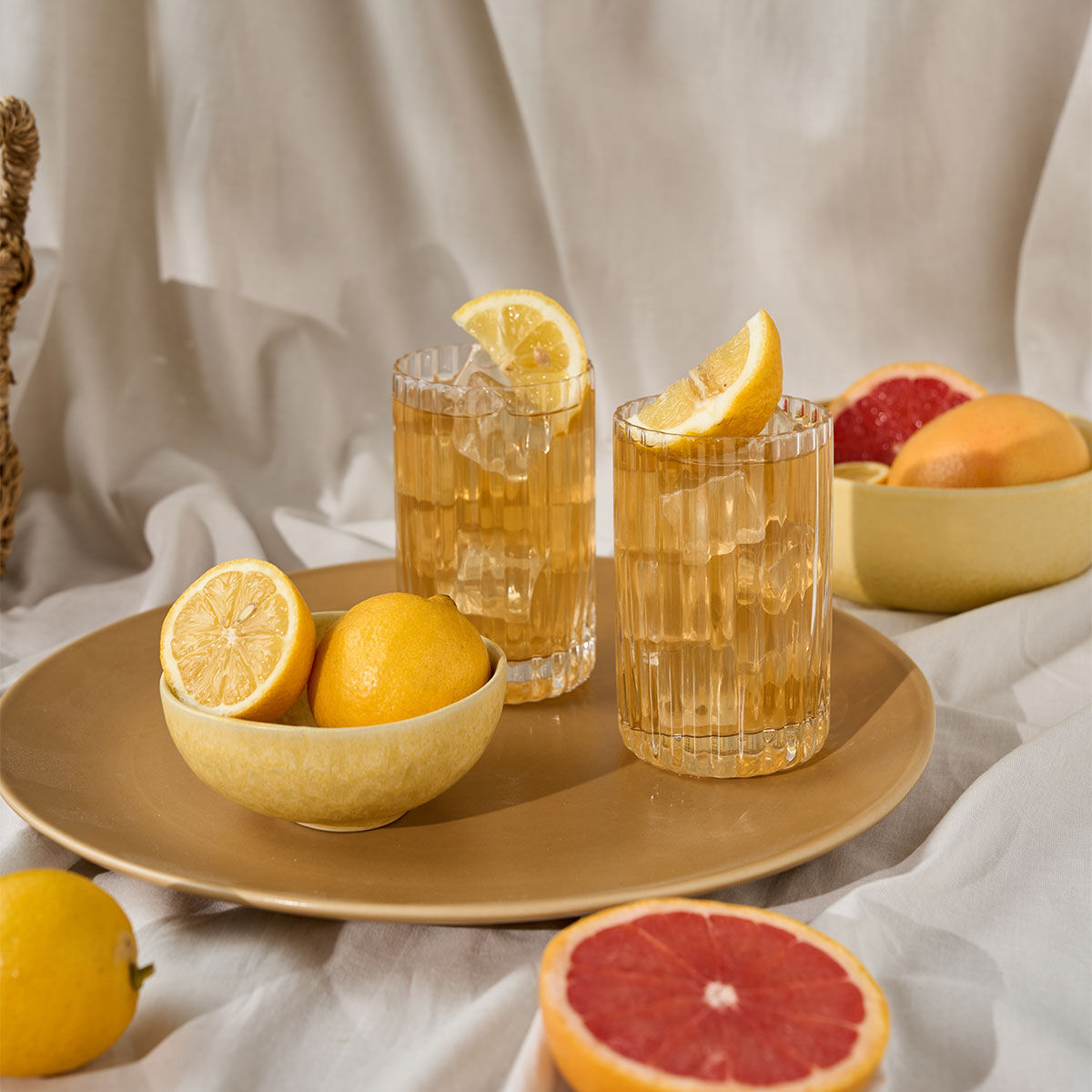 Zwei Gläser ORGANICS BIO Dankeschön Eistee mit Zitronenspalten auf einem goldenen Tablett, umgeben von Schalen mit Zitronen und halbierten Grapefruits, vor einem weißen Stoffhintergrund.