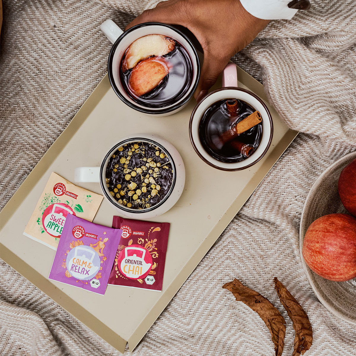 Eine Hand hält eine Tasse Tee mit Früchten neben zwei weiteren Tassen ORGANICS Kräutertee und Gewürztee auf einem Tablett mit Beuteln mit den Aufschriften „Sweet Apple“, „Calme & Relax“ und „Oriental Chai BIO“, die auf einer beigen Decke mit Äpfeln danebe