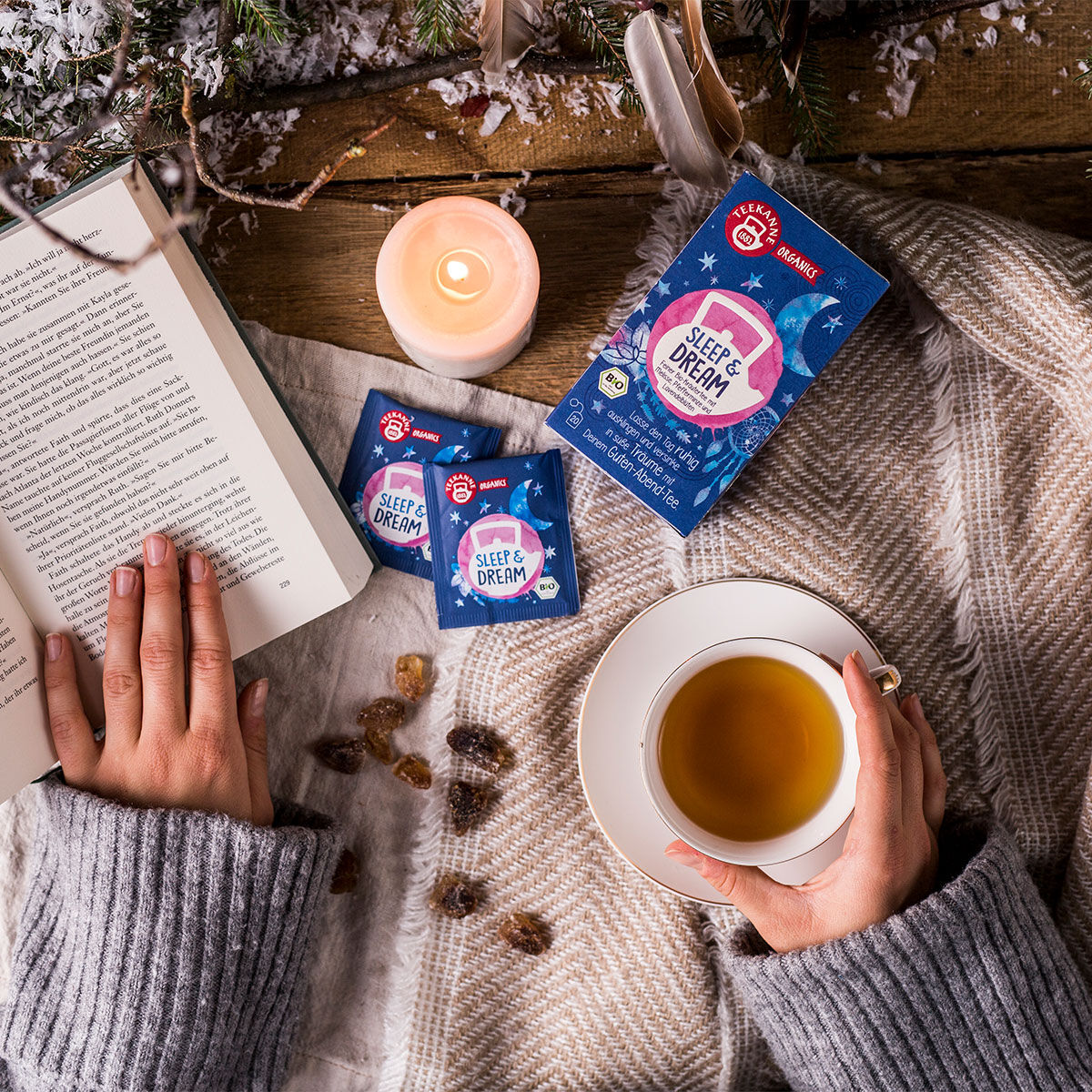 Eine Person im grauen Pullover liest ein Buch und hält dabei eine Tasse ORGANICS Sleep & Dream BIO Tee in der Hand. Teebeutel mit der Aufschrift Sleep & Dream und eine brennende Kerze liegen auf dem mit einer Decke bedeckten Holztisch und verbreiten eine 