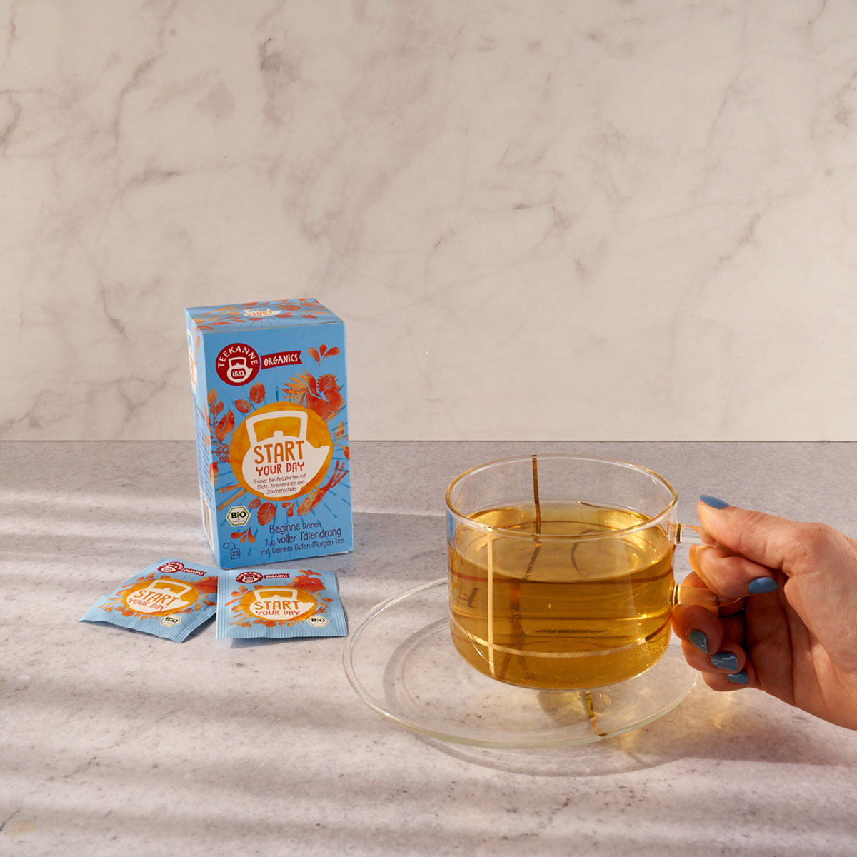 Eine Hand hält ein Glas mit hellem „Start Your Day BIO Kräutertee“ neben der Schachtel und zwei Beuteln auf einer hellen Arbeitsplatte mit Marmorhintergrund.
