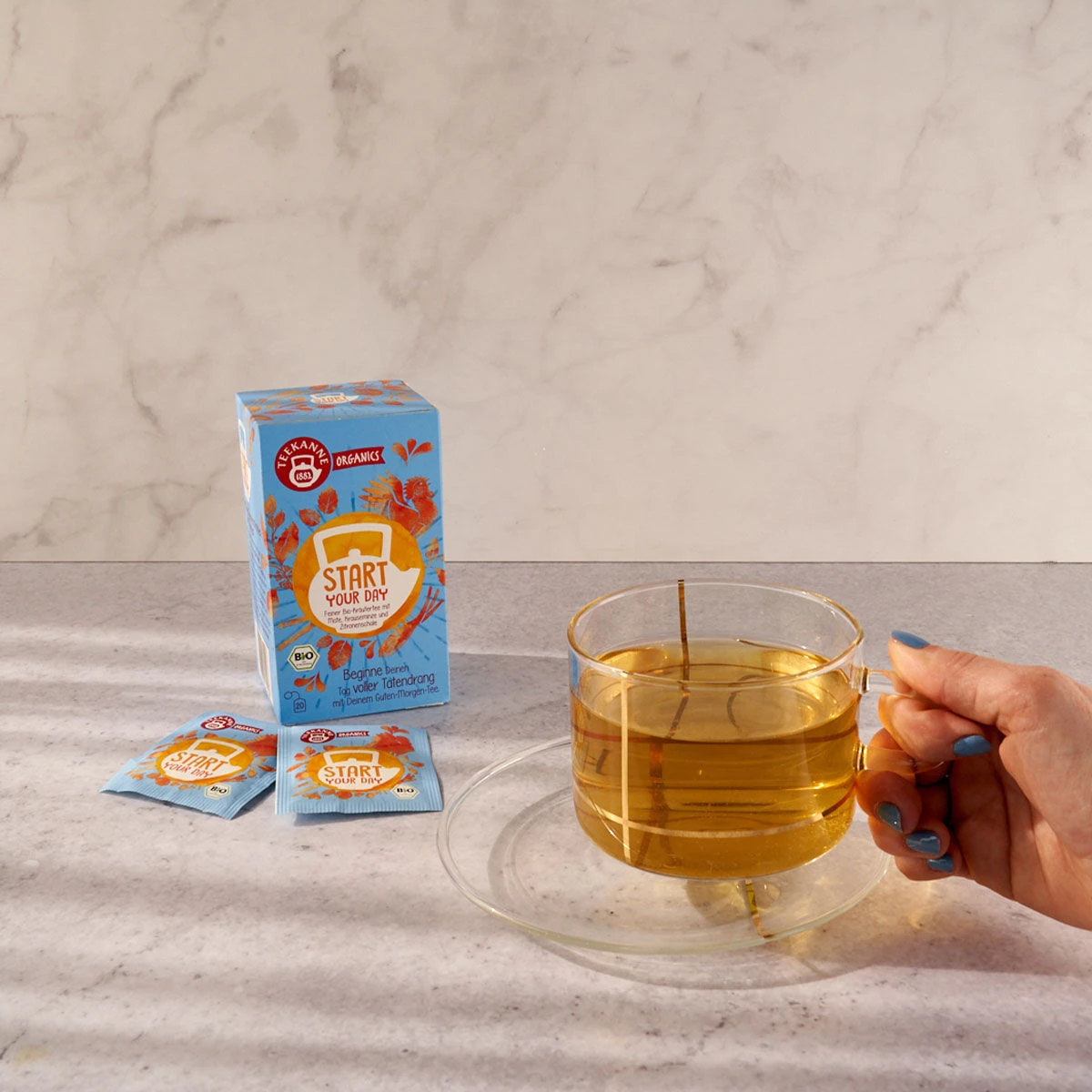 Eine Hand hält ein Glas mit hellem „Start Your Day BIO Kräutertee“ neben der Schachtel und zwei Beuteln auf einer hellen Arbeitsplatte mit Marmorhintergrund.