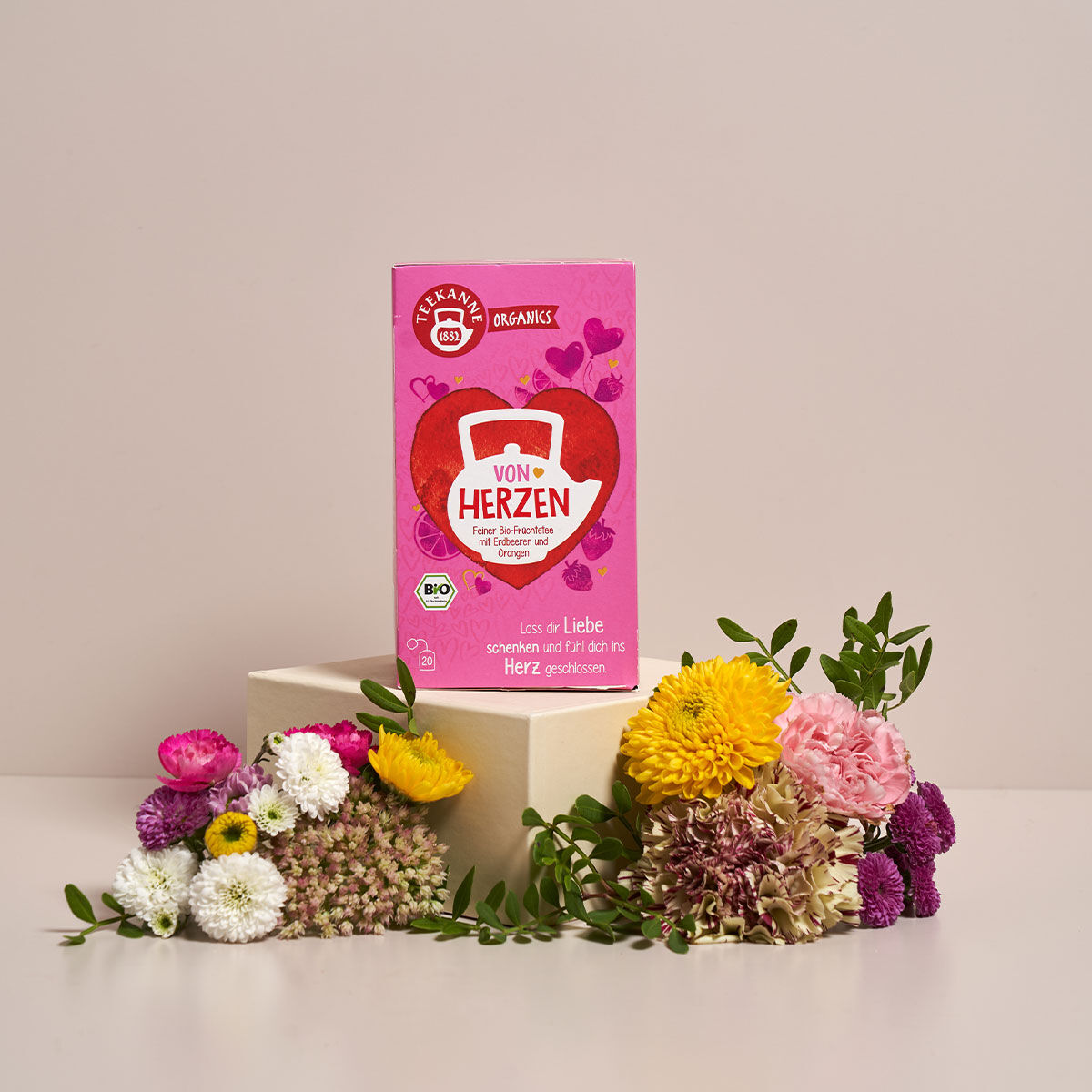 Eine rosa Schachtel ORGANICS BIO Von Herzen Tee steht auf einem beigen Podest, umgeben von bunten frischen Blumen, vor einem hellbeigen Hintergrund. Die Schachtel zeigt Herzmotive und deutschen Text.