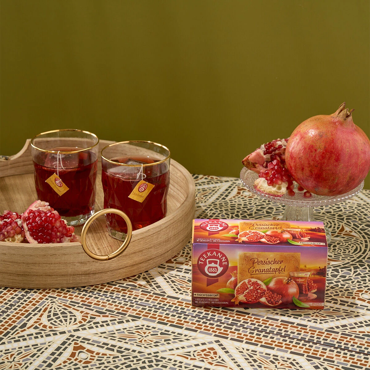 Auf einem gemusterten Tisch steht vor einem olivgrünen Hintergrund eine Schachtel Persischer Granatapfeltee mit zwei Gläsern rotem Tee, Granatapfelkernen, einem ganzen Granatapfel und einem Holztablett.