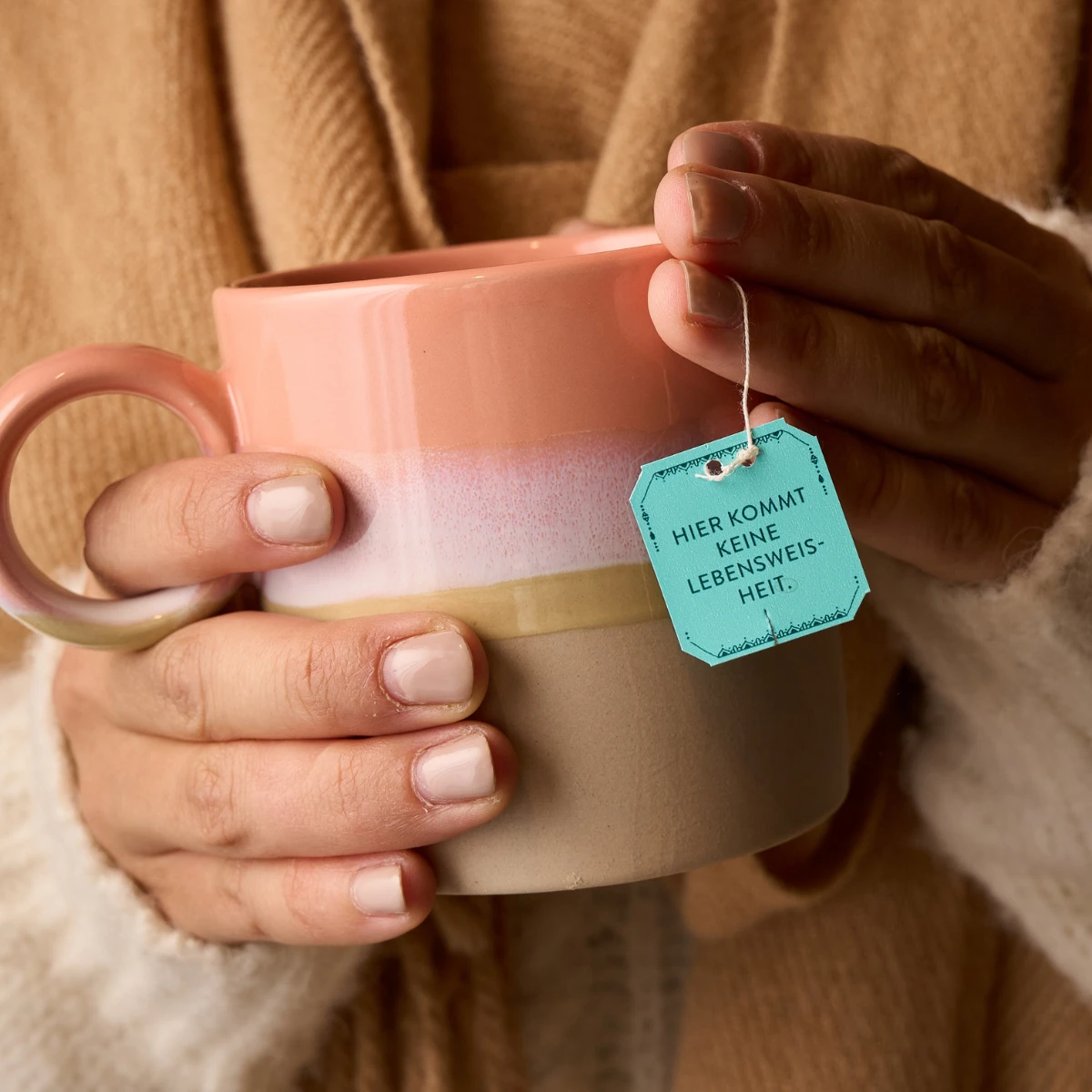 Eine Person in einem kuscheligen Pullover hält eine rosa-beige Tasse BIO NamasTee® Halsschmeichler mit einem blauen Teebeutelanhänger mit der Aufschrift „HIER KOMMT KEINE LEBENSWEISHEIT“ in der Hand.