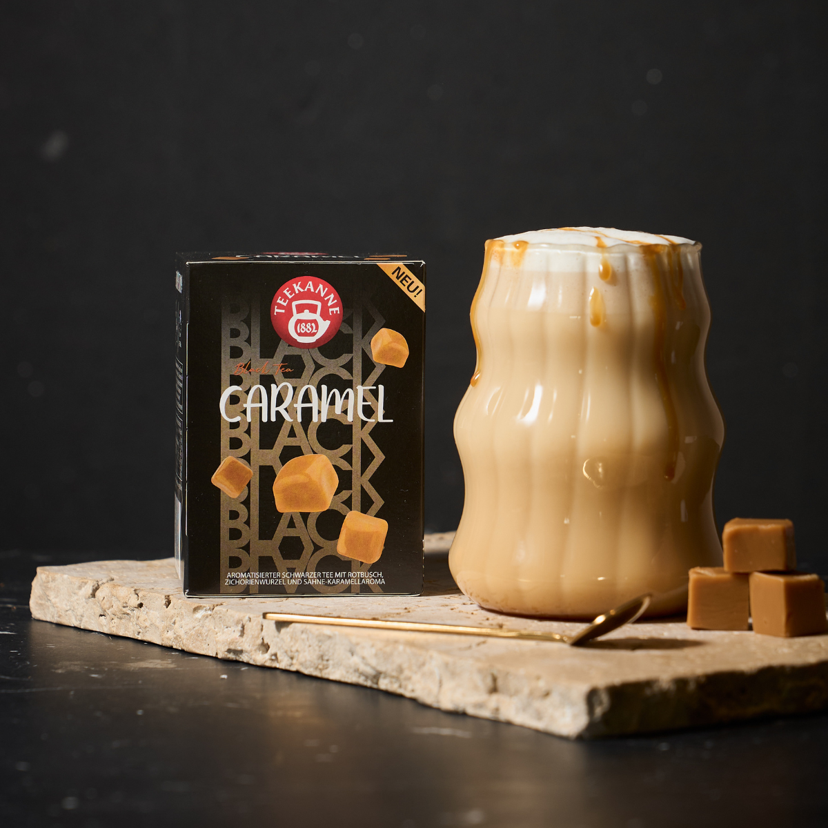 Ein gewelltes Glas Karamell-Latte mit aufgeschäumter Milch steht auf einer Steinplatte neben einer Schachtel Black Tea Caramel und Karamellbonbons. Der dunkle Hintergrund hebt das cremige Getränk, die Verpackung und das reichhaltige Aroma des Schwarztees 