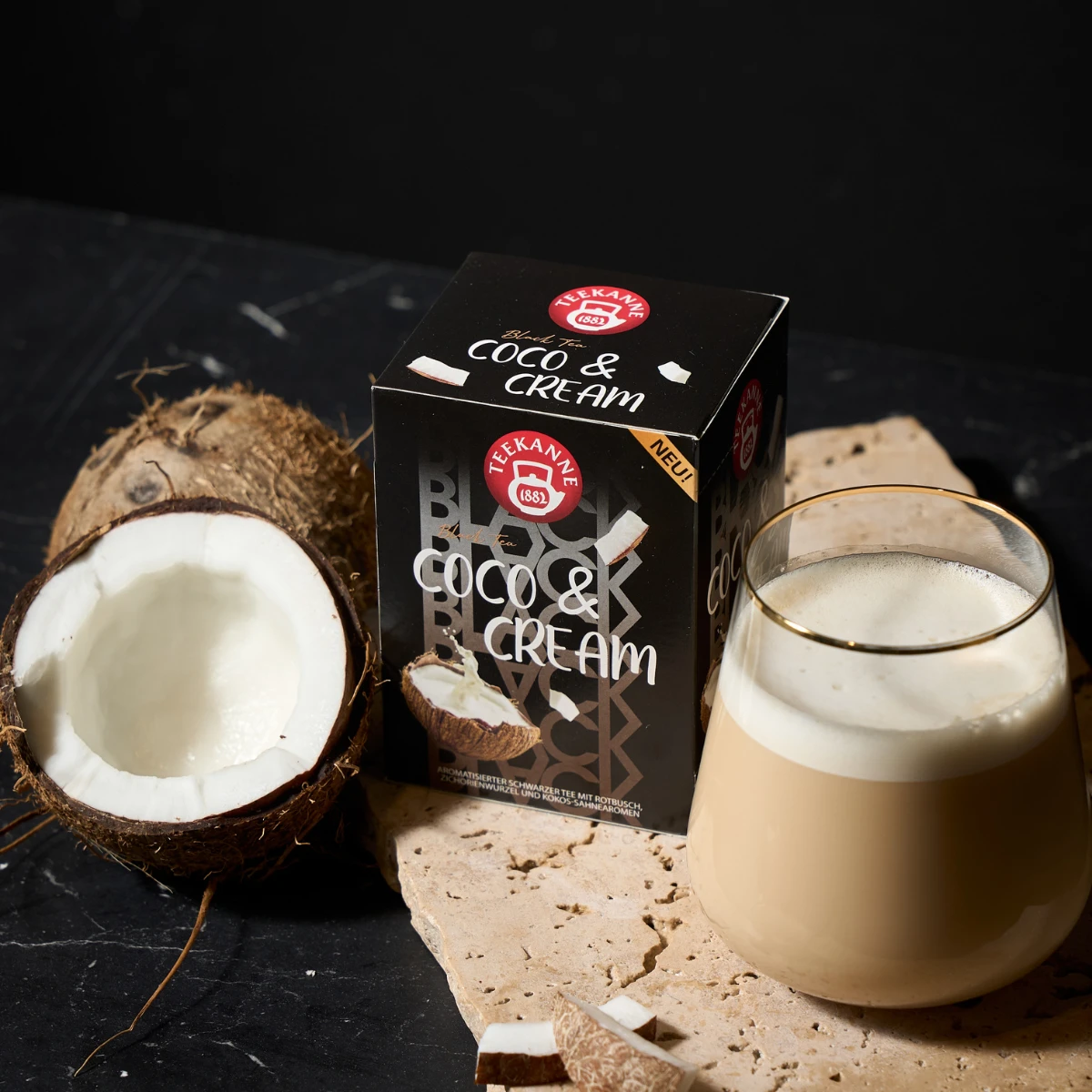 Ein Glas cremiges Kokosnussgetränk steht neben einer Schachtel mit der Aufschrift „Black Tea Coco & Cream“, einer halbierten Kokosnuss und Kokosnussstücken auf einer dunklen Oberfläche mit einer beigen Matte.