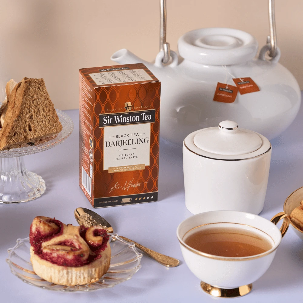 Eine Schachtel Sir Winston Darjeeling steht auf einem Tisch mit einer Teekanne, einer Zuckerdose, einer mit Tee gefüllten Teetasse und Tellern mit einem Stück Kuchen und einer Obsttorte.