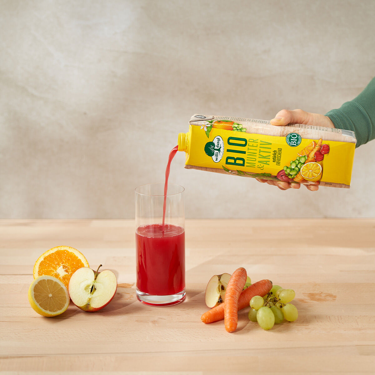 Eine Hand gießt Munter & Aktiv Bio aus einem gelben Karton in ein Glas. Auf dem Tisch stehen eine Orangenhälfte, Apfelscheiben, Zitrone, Karotten und grüne Trauben. Der Hintergrund ist neutral und hell.