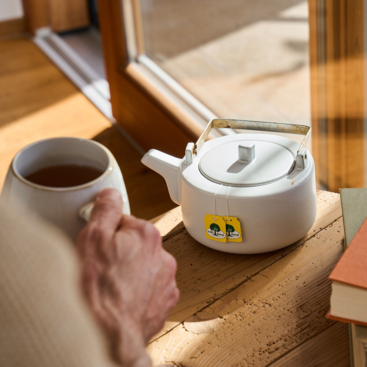 Eine Person genießt eine Tasse Tee an einem Holztisch mit einer weißen Teekanne, zwei gelben Teebeutelanhängern und Büchern in der Nähe – ein idealer gemütlicher Moment, um mit Ihrem Schlaf-Freund BIO neben einem sonnenbeschienenen Fenster zu entspannen.
