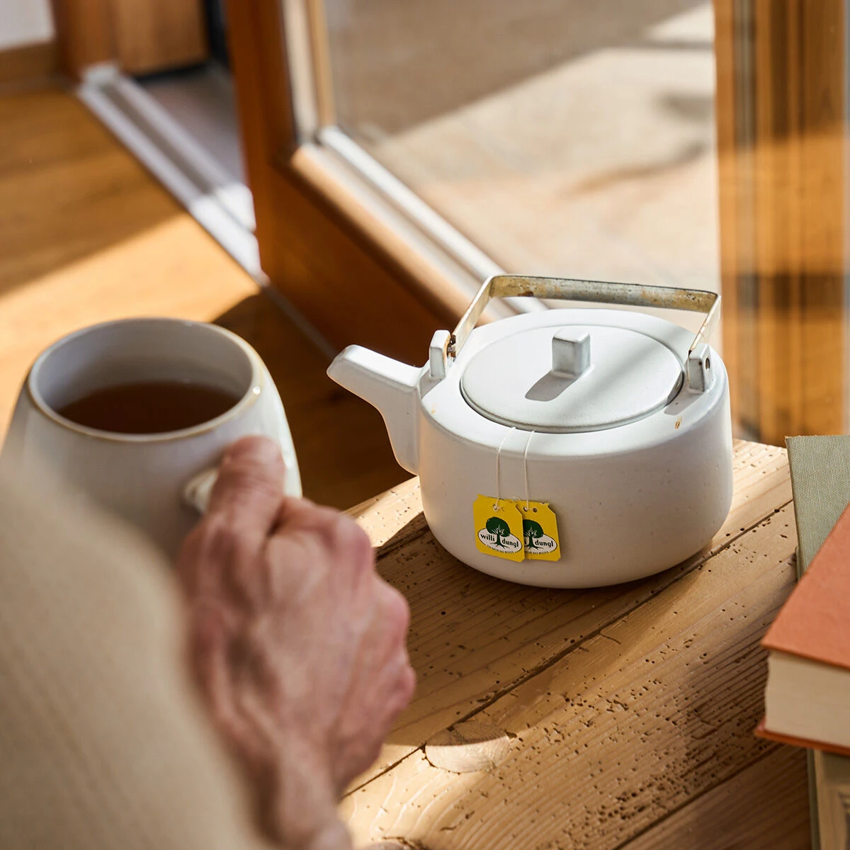 Eine Person genießt eine Tasse Tee an einem Holztisch mit einer weißen Teekanne, zwei gelben Teebeutelanhängern und Büchern in der Nähe – ein idealer gemütlicher Moment, um mit Ihrem Schlaf-Freund BIO neben einem sonnenbeschienenen Fenster zu entspannen.