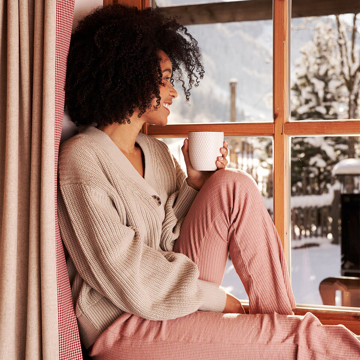 Eine Frau mit lockigem Haar sitzt lächelnd am Fenster und hält eine Tasse Bio Winter Orange in der Hand. Sie trägt einen beigen Pullover und eine rosa Hose. Vor dem Fenster sind verschneite Bäume und Berge zu sehen, während warmes Sonnenlicht den gemütlic