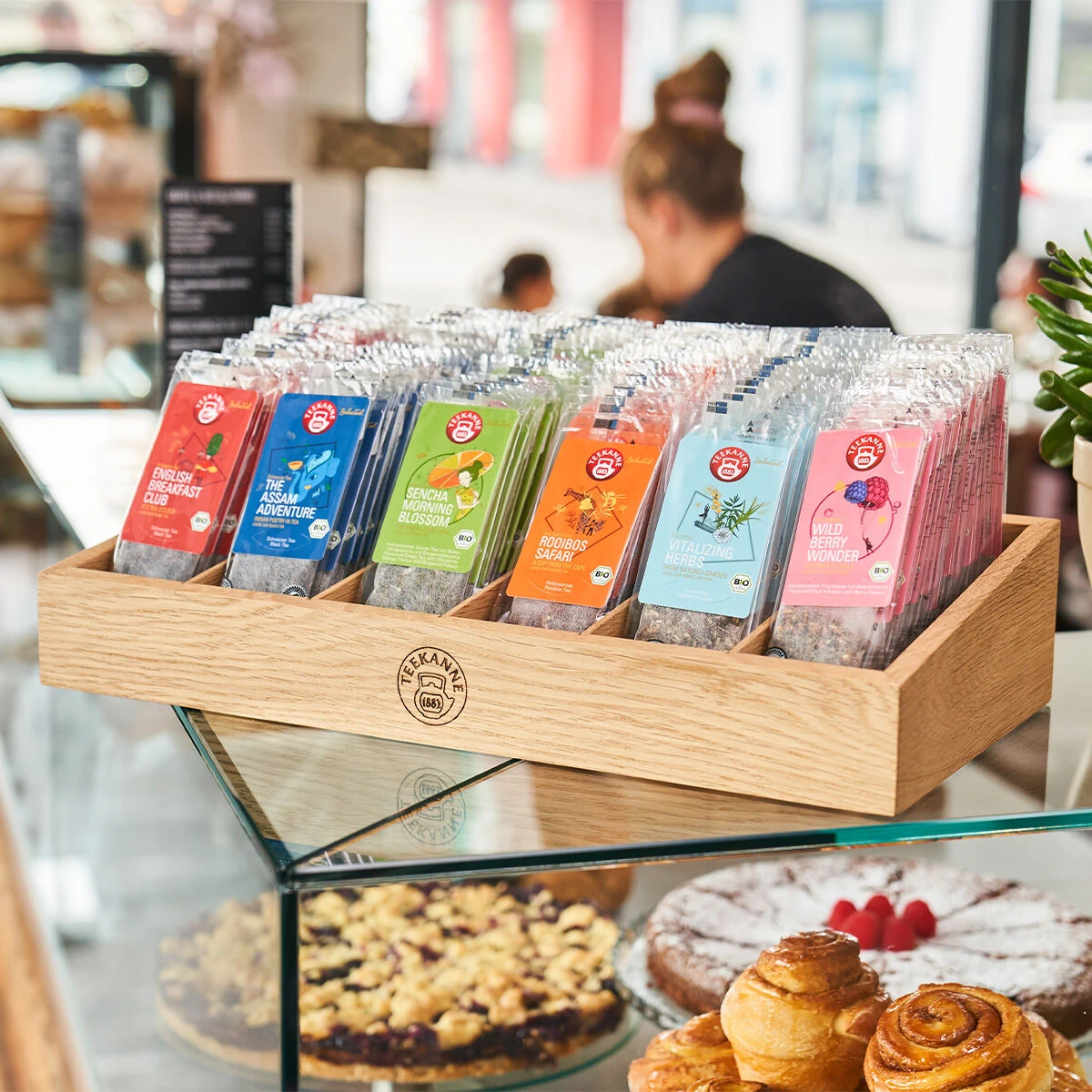 Eine Holzvitrine auf einer Glastheke präsentiert bunte Schokoriegel, während verschiedene Gebäcke und Kuchen neben der Vitalizing Herbs Luxury Bag liegen. Im Hintergrund des hellen Cafés ist eine Person unscharf zu erkennen.