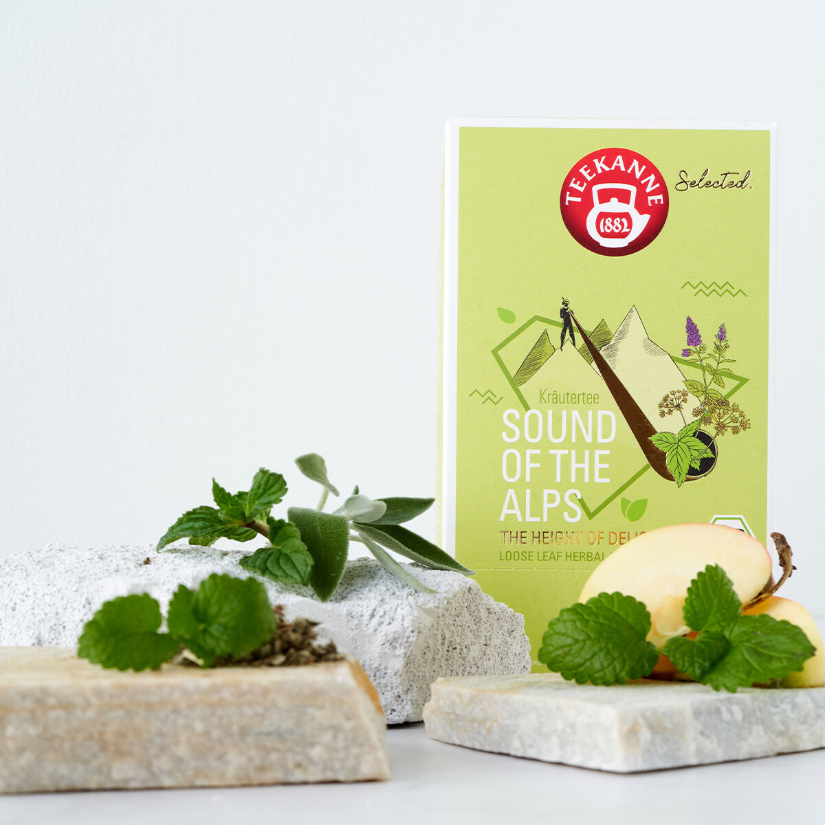 Eine Schachtel Sound of the Alps Luxury Cup BIO steht aufrecht, umgeben von frischen Kräutern, Apfelscheiben und einer Steinplatte auf einem schlichten weißen Hintergrund.