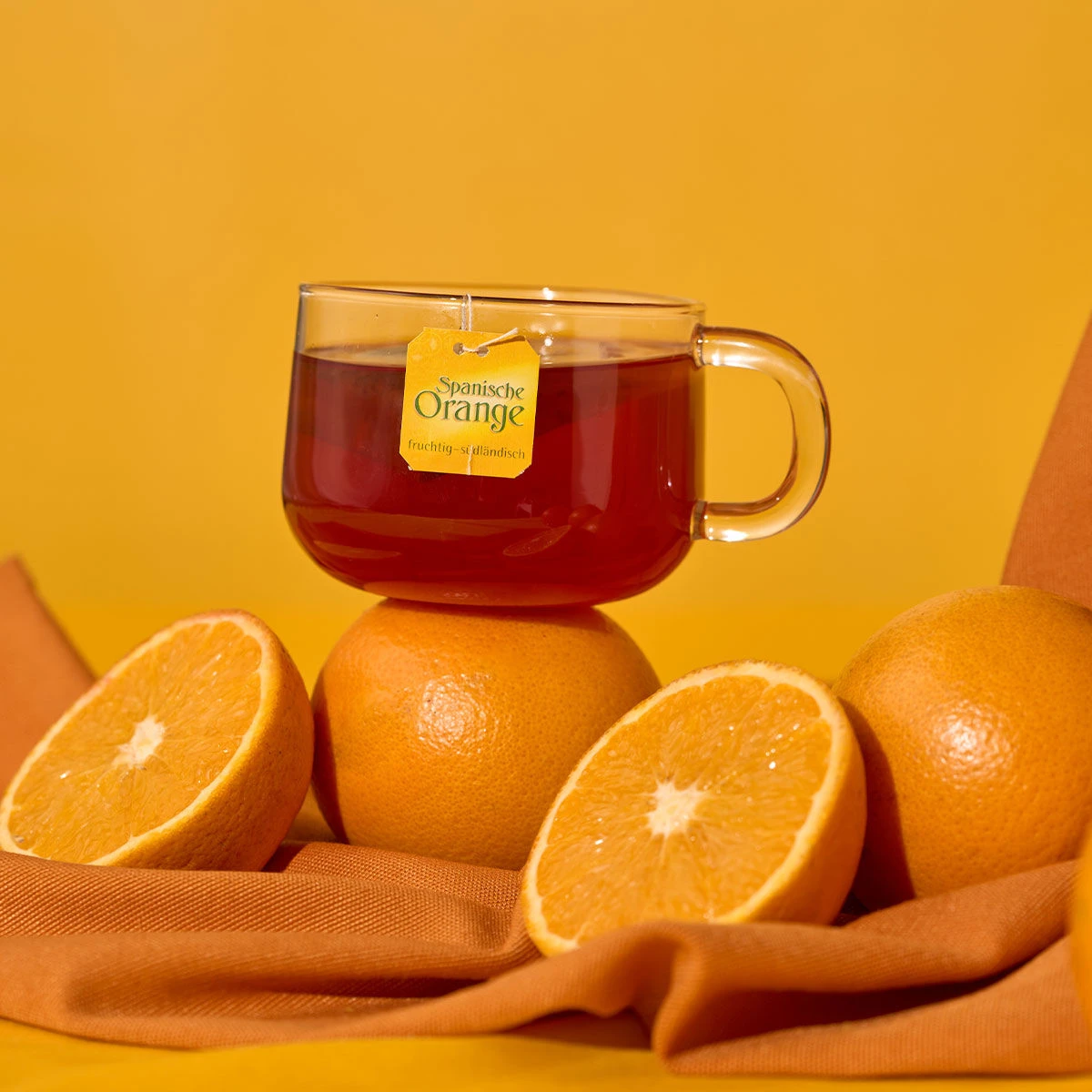 Eine Glastasse Spanischer Orangentee mit einem Etikett mit der Aufschrift „Spanische Orange“ steht auf einer Orange, umgeben von ganzen und halbierten Orangen auf einem orangefarbenen Tuch, alles vor einem warmen orangefarbenen Hintergrund.
