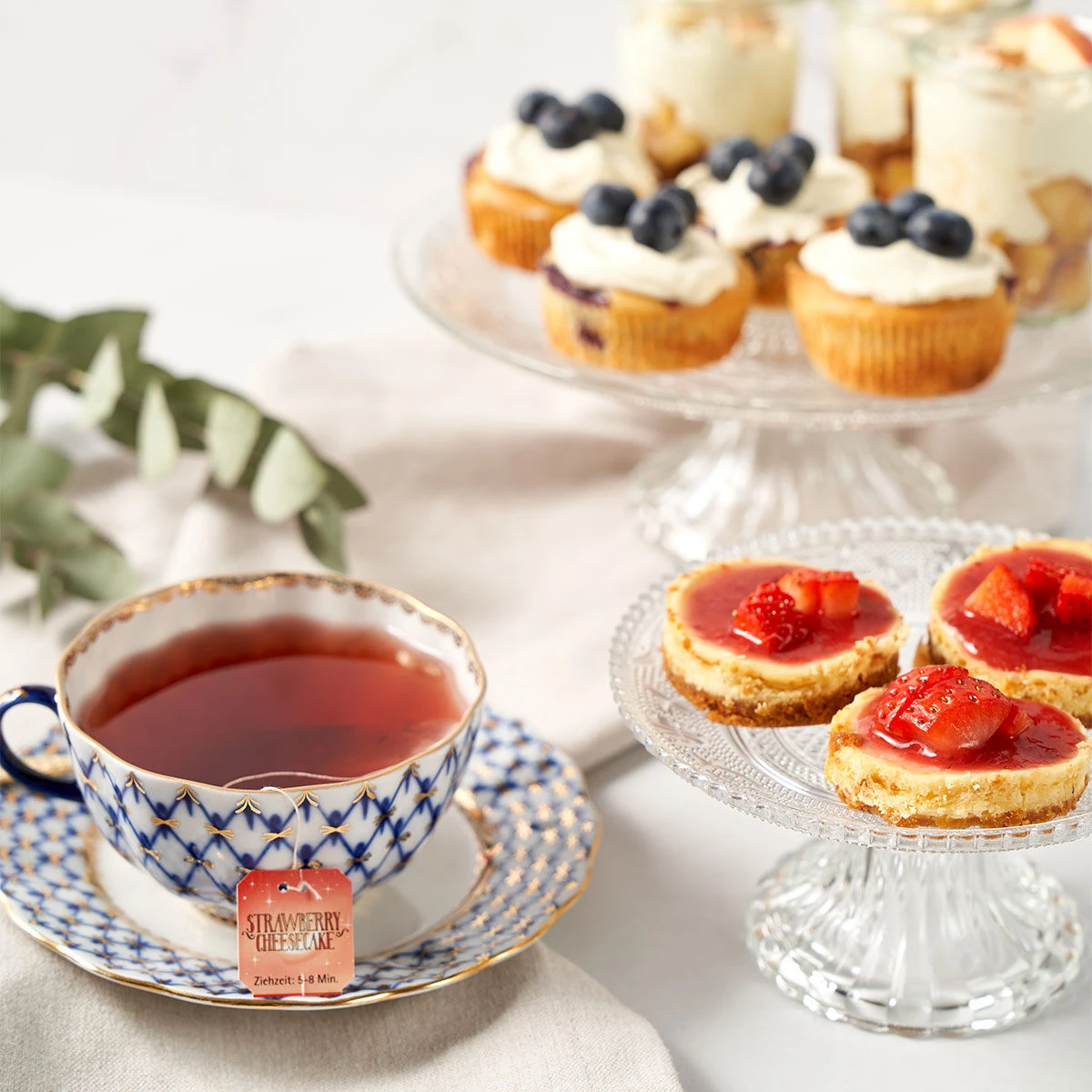 Eine gemusterte Tasse mit roten Sweeteas steht neben Erdbeerkäsekuchen mit Erdbeeren und Mini-Cupcakes mit Sahne und Blaubeeren, alles hübsch auf Glasständern präsentiert.