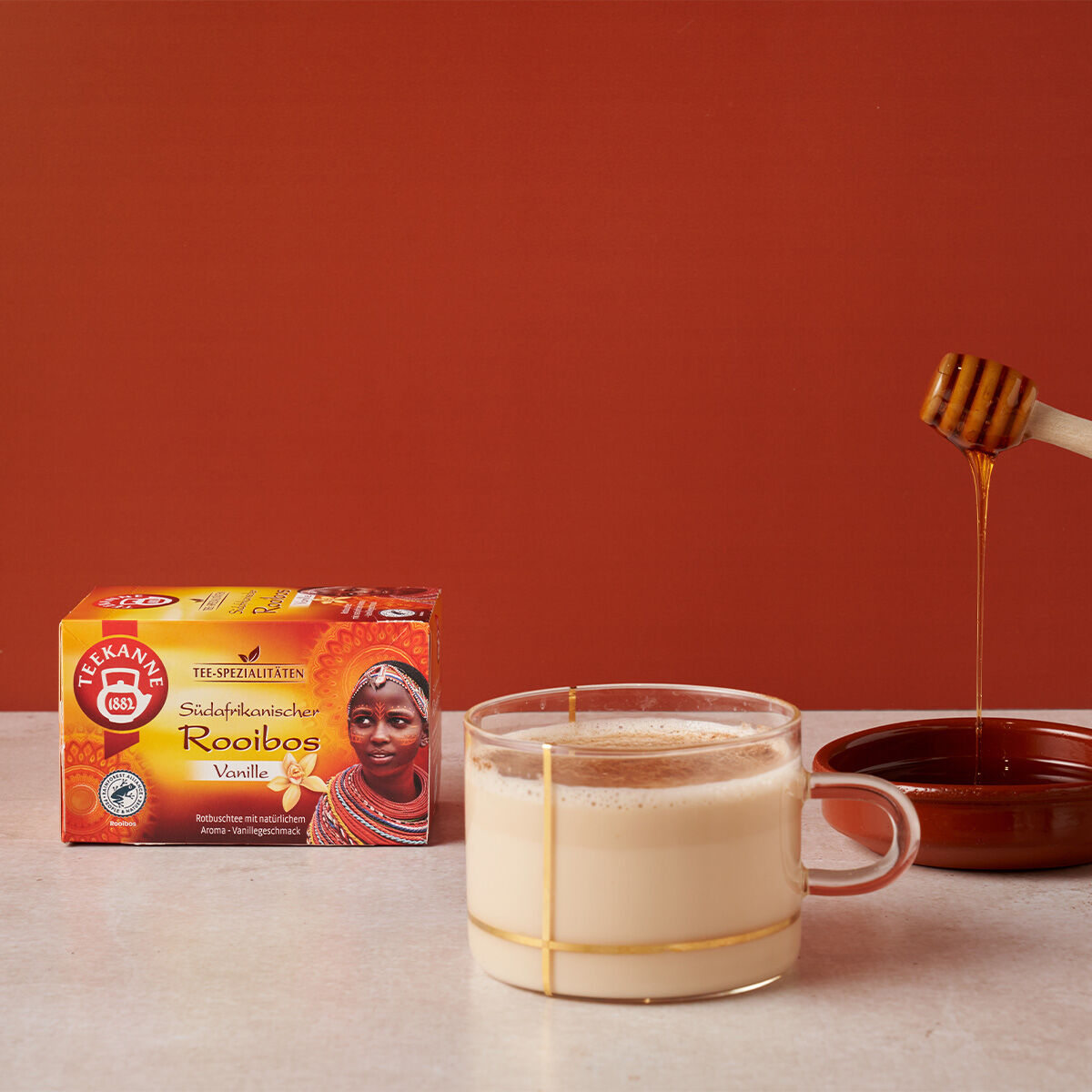 Eine Tasse cremigen Rooibos-Tees steht neben Honig und einem Schöpflöffel. Dahinter präsentiert eine Schachtel Südafrikanischer Rooibos Vanille RFA seine koffeinfreien und erfrischenden Eigenschaften vor einem warmen roten Hintergrund.