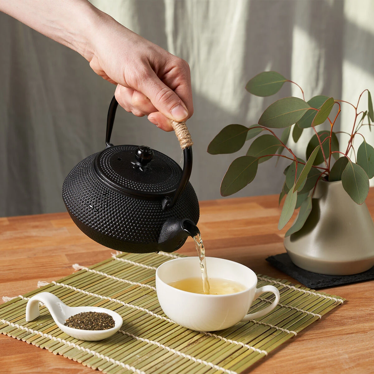 Eine Hand gießt Teefix Grüner Tee aus einer schwarzen Teekanne in eine weiße Tasse auf einer Bambusmatte, während ein Löffel mit frischen grünen Teeblättern und eine Vase mit Grünzeug im Hintergrund für eine authentische Note sorgen.