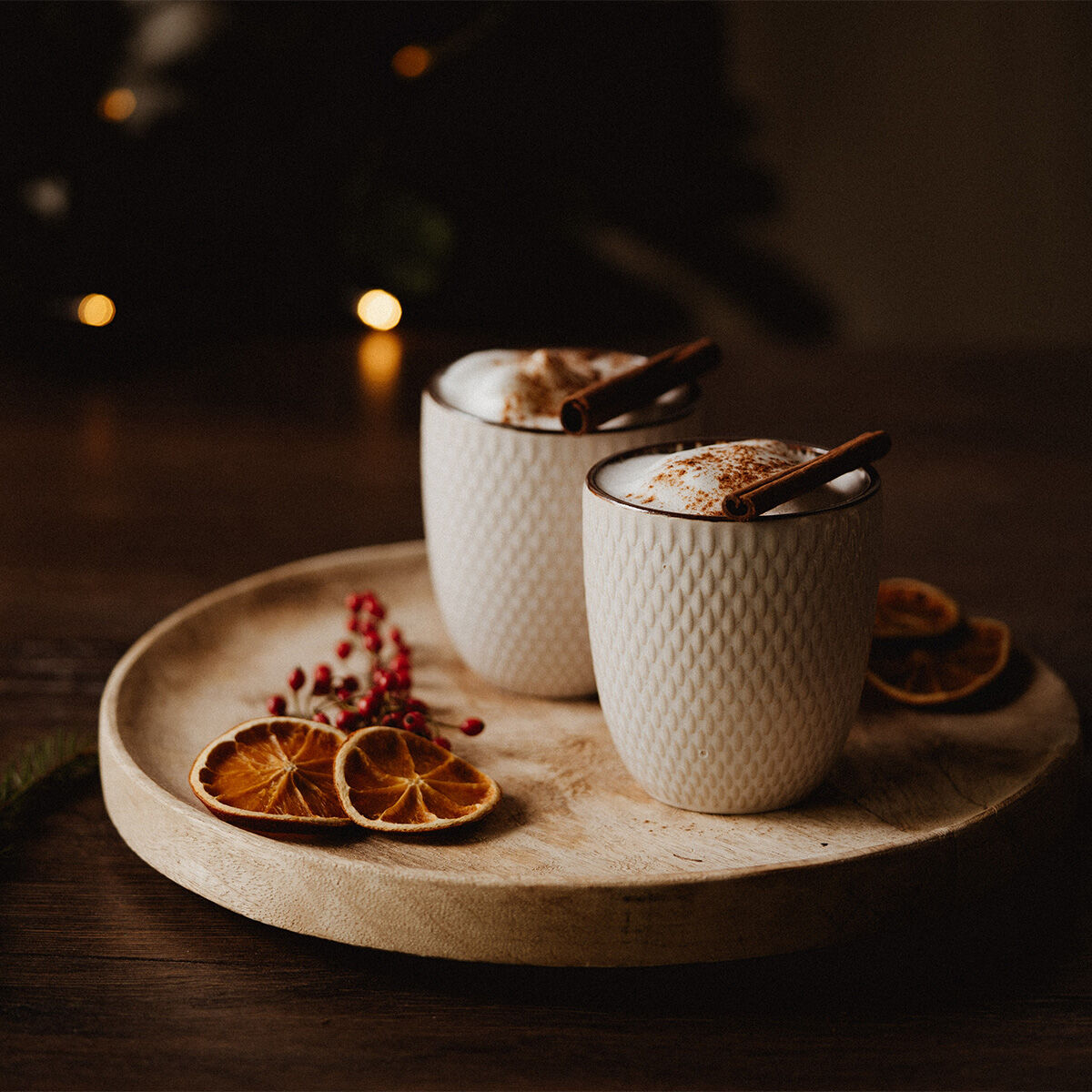Zwei weiße Becher gefüllt mit schaumiger Trinkschokolade, garniert mit Zimtstangen, stehen auf einem Holztablett mit getrockneten Orangenscheiben und roten Beeren vor einem gemütlichen dunklen Hintergrund.