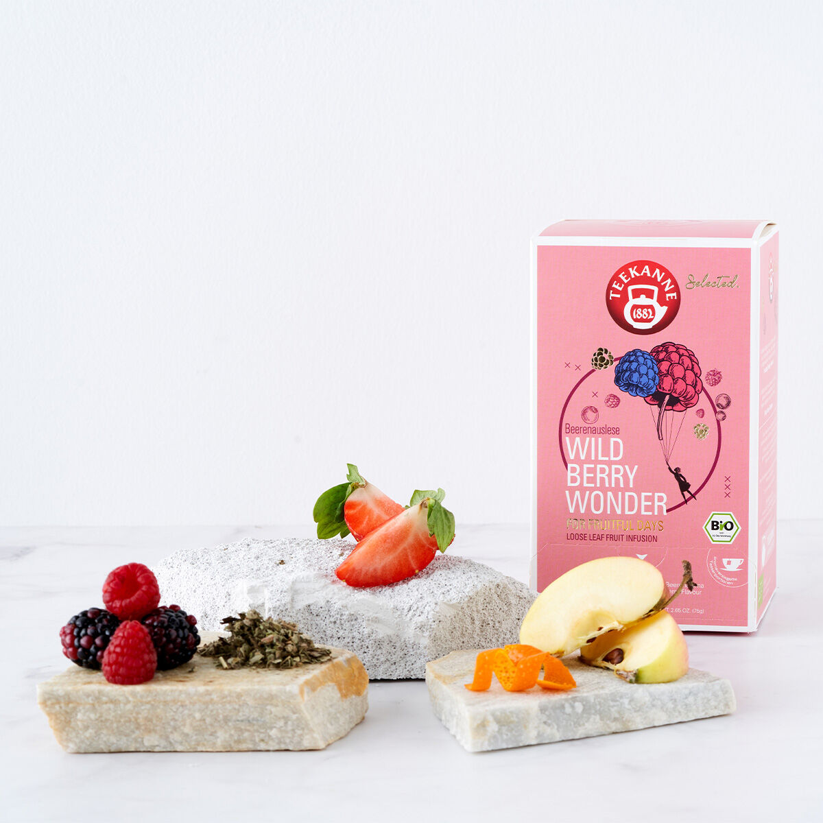 Eine Schachtel Wild Berry Wonder Luxury Cup BIO steht hinter Steinplatten, die mit frischen Erdbeeren, Beeren, Apfelscheiben, Orangenschale und losem Tee auf weißem Hintergrund belegt sind.