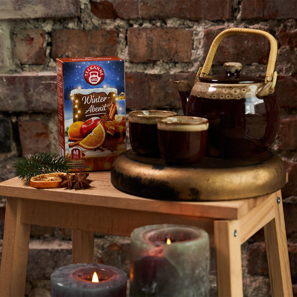 Eine rustikale Szene mit einer Schachtel Winterabendtee, einer braunen Teekanne, zwei Tassen, Kerzen und getrockneten Orangenscheiben auf einem Holzhocker vor einer Ziegelwand.