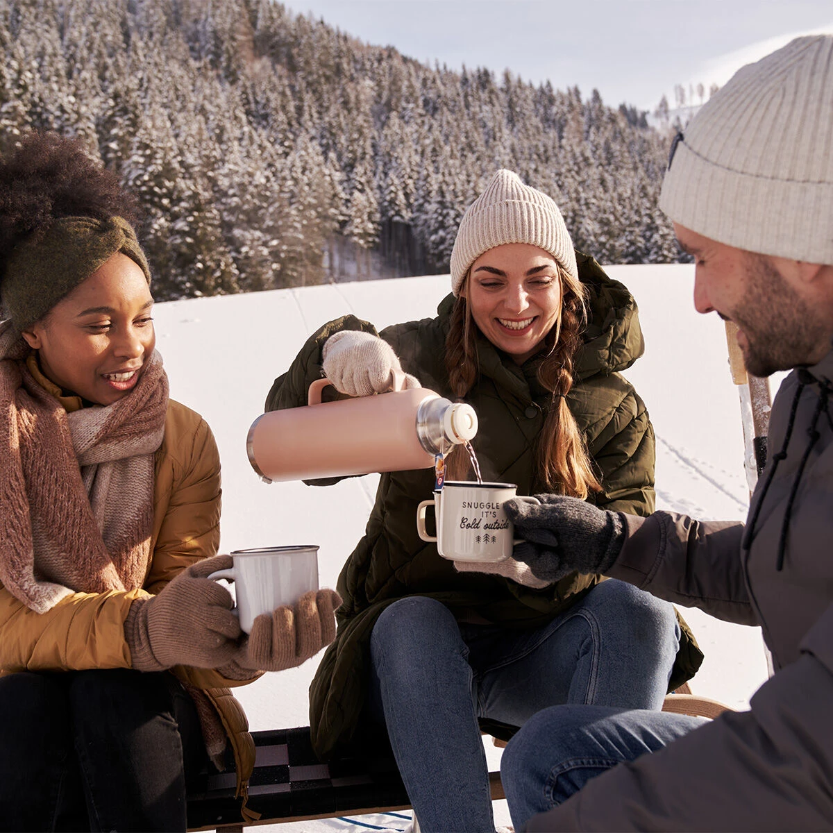 Drei Freunde in gemütlicher Winterkleidung lächeln sich an einem verschneiten Hang an, während sie Winterzauber-Tee aus einer Thermoskanne in Tassen gießen, umgeben von schneebedeckten Bäumen und Bergen – ein wahrhaft magischer Moment.