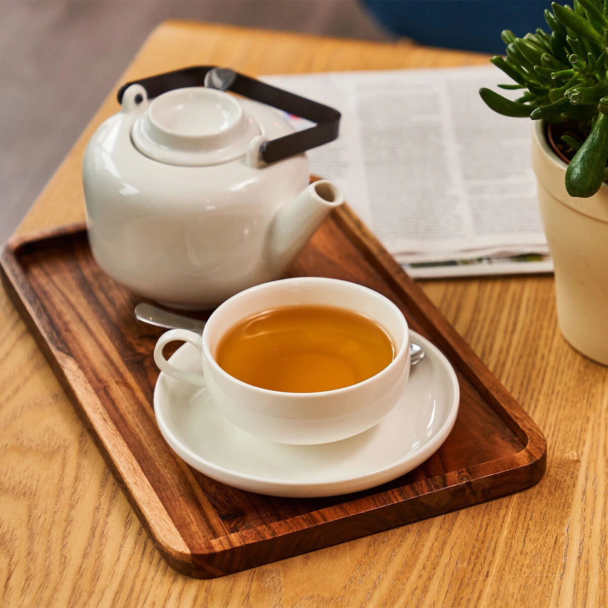 Ein 8-l-Heißwassergerät gibt heißes Wasser für Tee in eine weiße TEEKANNE-Teekanne und -Teetasse auf einem Tablett auf einem Holztisch aus, ergänzt durch stilvolles Zubehör, einen silbernen Löffel auf der Untertasse und ein offenes Magazin für ein einlade