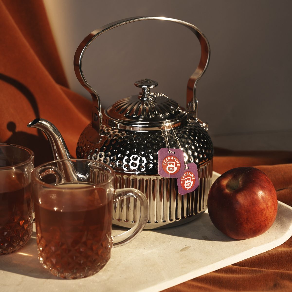 Eine silberne Teekanne mit zwei Teeetiketten steht auf einem hellen Tablett neben einem Türkischen Apfel und zwei mit Früchtetee gefüllten Glasbechern, alles vor einem warmen braunen Stoffhintergrund.