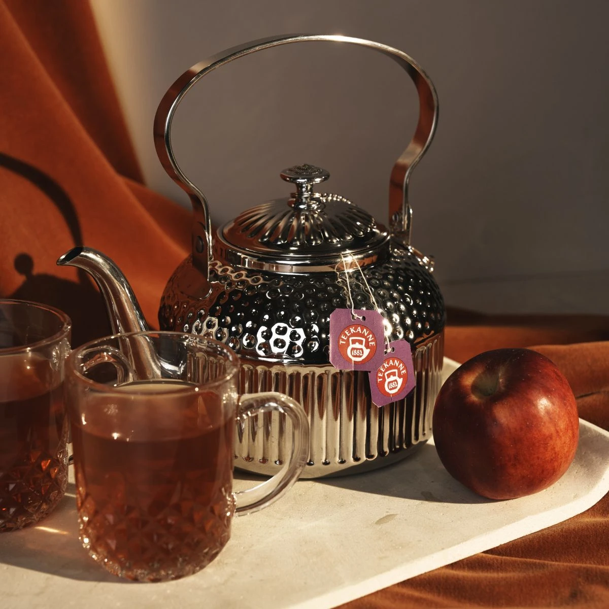 Eine silberne Teekanne mit zwei Teeetiketten steht auf einem hellen Tablett neben einem Türkischen Apfel und zwei mit Früchtetee gefüllten Glasbechern, alles vor einem warmen braunen Stoffhintergrund.