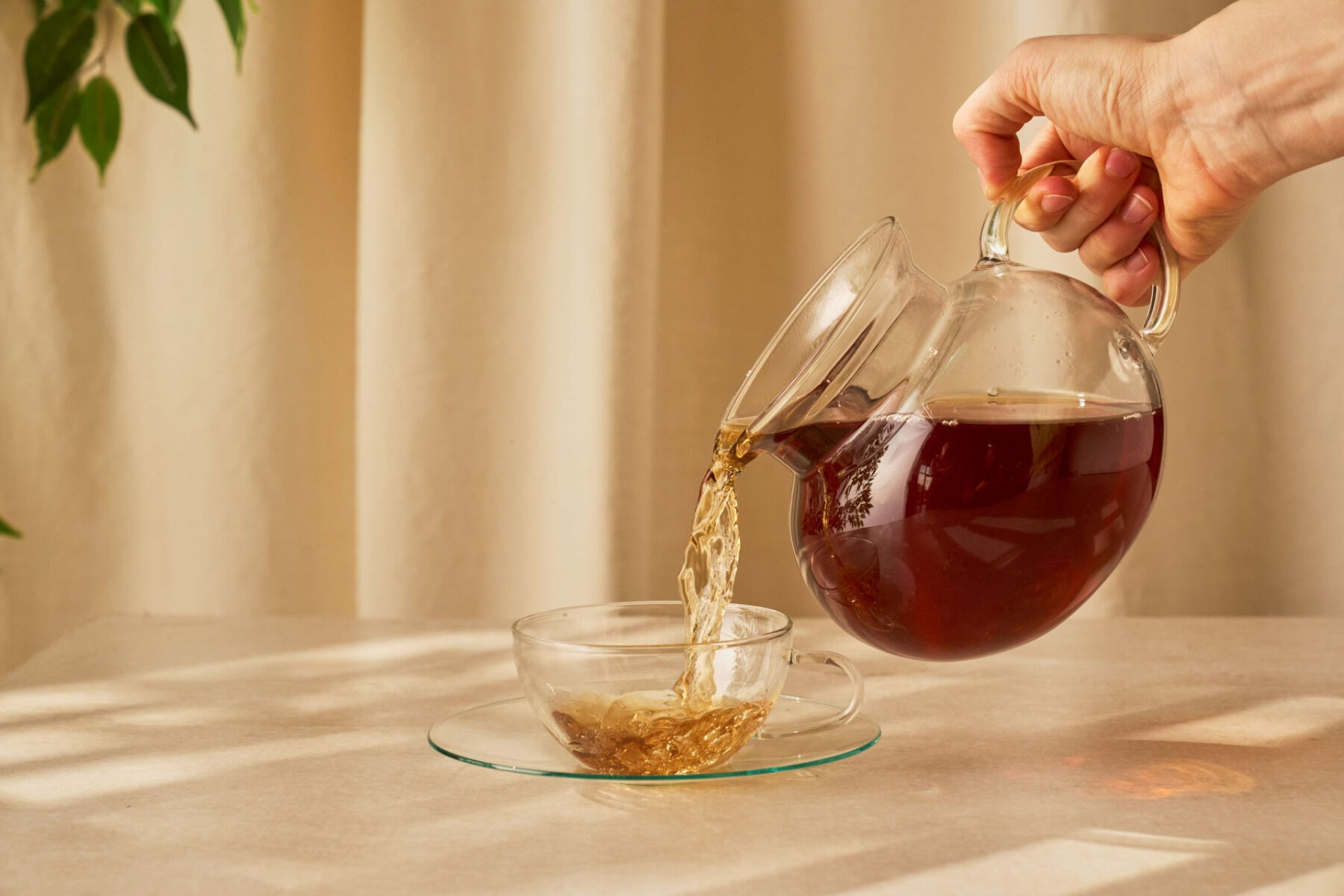 Hand gießt Tee aus einer gläsernen Teekanne in eine kleine gläserne Tasse auf einem hellen Tisch