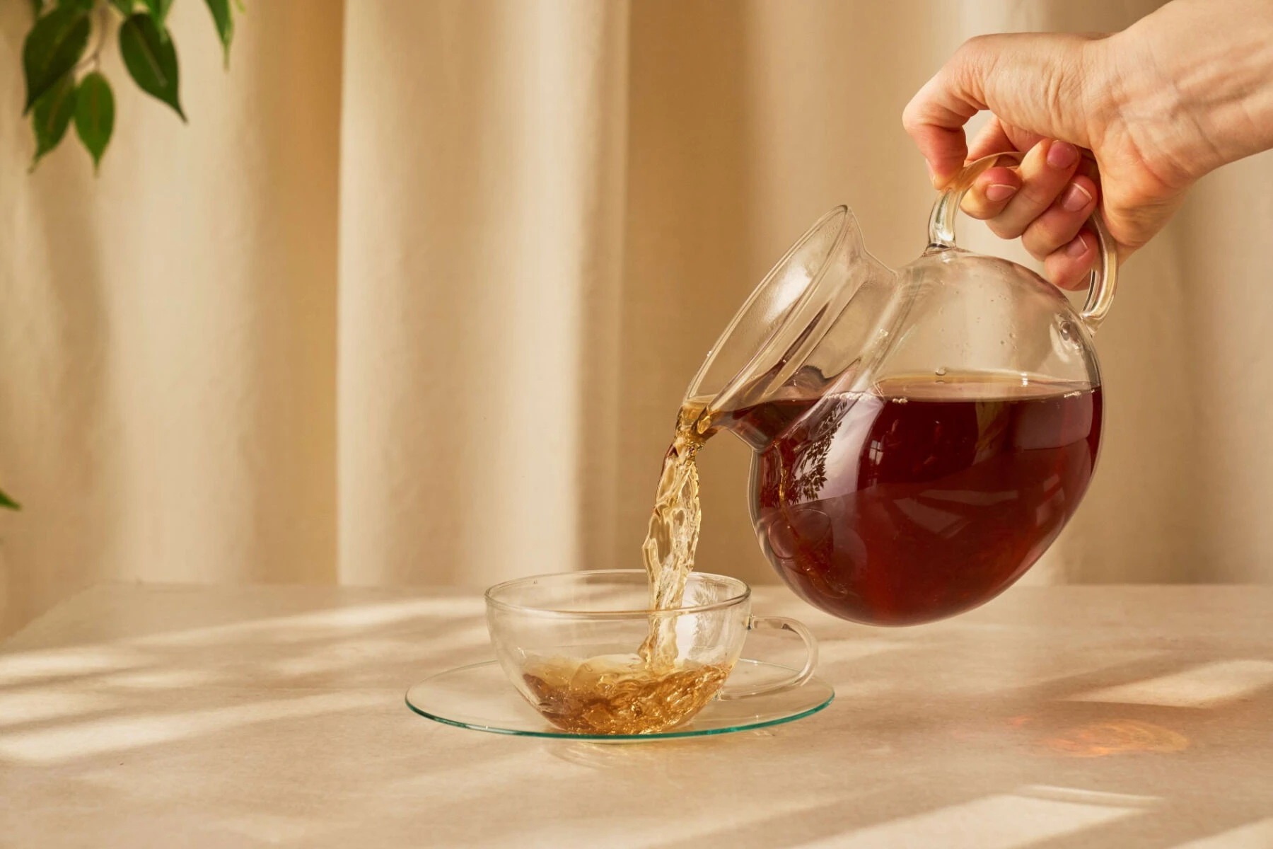 Hand gießt Tee aus einer gläsernen Teekanne in eine kleine gläserne Tasse auf einem hellen Tisch