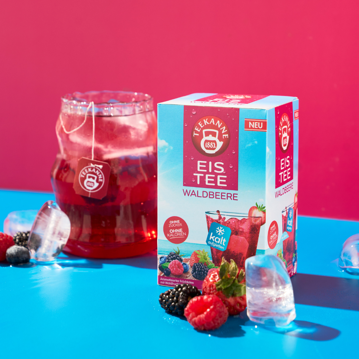 Eine Schachtel Waldbeeren steht neben einem Glas rotem Eistee mit Eiswürfeln, umgeben von verschiedenen Beeren auf einer leuchtend blauen Oberfläche und verspricht pure Erfrischung vor einem rosa Hintergrund.
