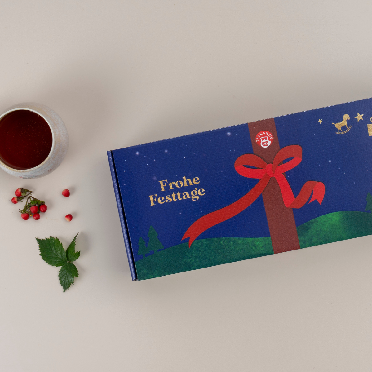 Die Wintertee-Geschenkbox, eine festliche blaue Schachtel mit roter Schleife und dem Text „Frohe Festtage“, steht auf einer hellen Oberfläche neben einer Tasse Tee, roten Beeren und grünen Blättern.