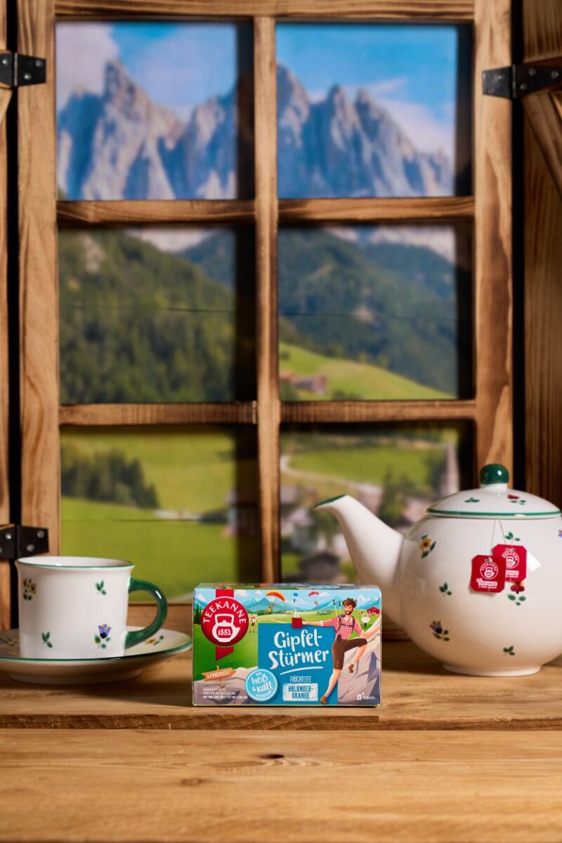 Eine weiße Teekanne, eine passende Tasse und eine Schachtel Gipfelstürmer-Tee stehen auf einem Holztisch an einem Fenster mit Blick auf die Berge.